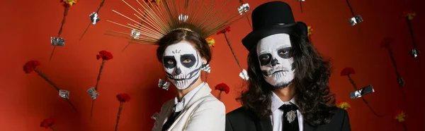 Couple en dia de los muertos maquillage et tenue de fête posant sur fond rouge avec des fleurs, bannière — Photo de stock