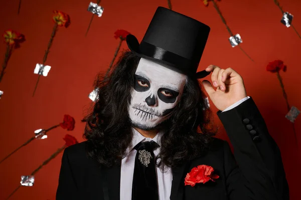 Hombre extravagante en el maquillaje del cráneo tocando sombrero de copa sobre fondo rojo con claveles, Día de Muertos - foto de stock