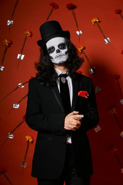 Man in skull makeup and festive attire looking at camera on red floral backdrop, dia de los muertos — Stock Photo