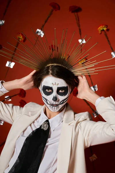Женщина в сахарном черепе макияж и белый костюм регулируя праздничную корону на красном фоне с цветами — стоковое фото