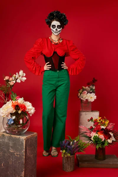 Woman in sugar skull makeup near traditional dia de los muertos ofrenda with bright flowers on red — Stock Photo