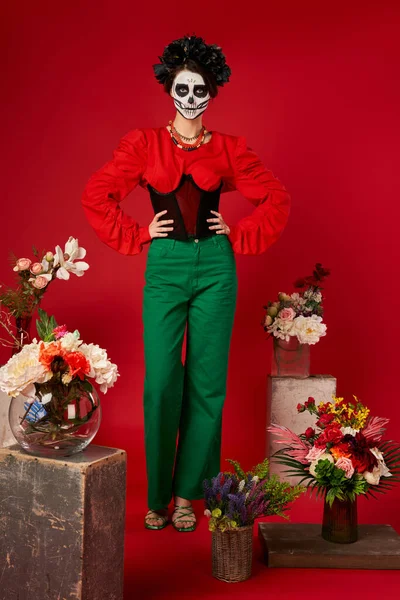 Mujer en maquillaje de cráneo de azúcar cerca del tradicional día de los muertos ofrenda con flores brillantes sobre rojo - foto de stock