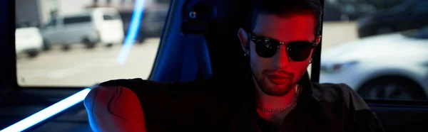 Young good looking man in sunglasses with tattoo on arm posing in his car, fashion concept, banner — Stock Photo