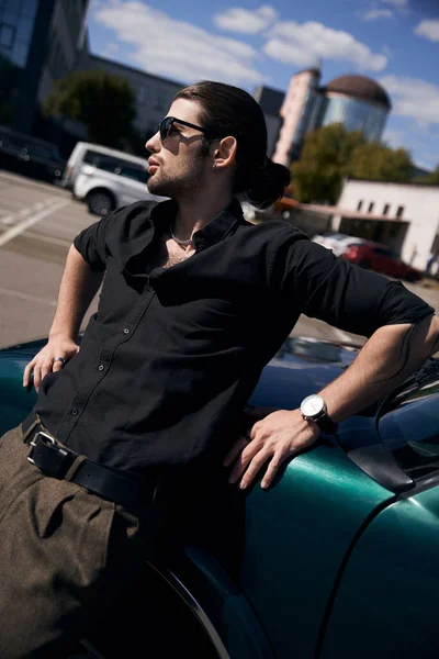Vertikale Aufnahme eines hübschen, verführerischen Fahrers in schwarzem Freizeitoutfit, der in der Nähe des Autos posiert und wegschaut — Stockfoto