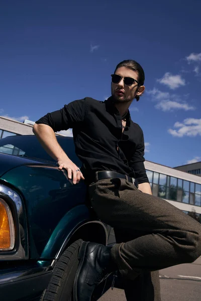 Hombre barbudo con estilo con tatuaje y gafas de sol en traje negro de pie junto a su coche, conductor sexy - foto de stock