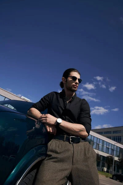 Élégant modèle masculin sexy avec queue de cheval en tenue décontractée noire posant à côté de sa voiture, en plein air — Photo de stock