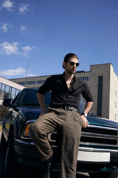 Joven hombre con estilo en traje casual negro de pie junto a su coche verde y mirando hacia otro lado, la moda - foto de stock