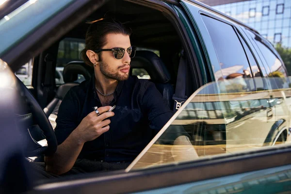 Atractivo hombre elegante con cola de caballo y gafas de sol en traje casual negro fumar cigarrillo en el coche - foto de stock