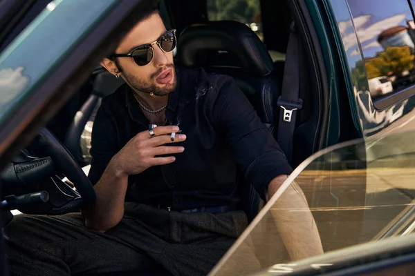Sexy modèle masculin en chemise noire avec des lunettes de soleil derrière volant et cigarette fumante — Photo de stock