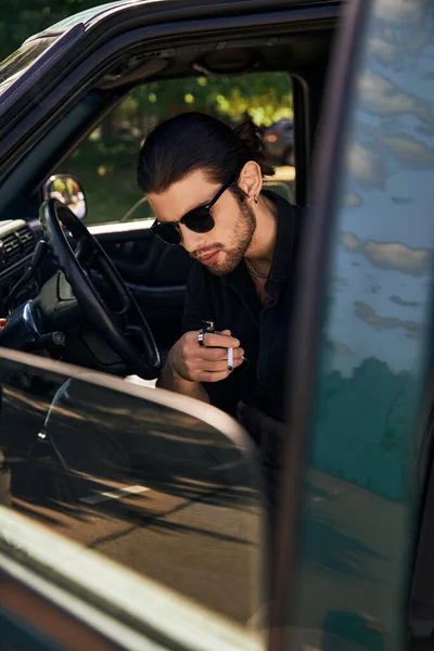 Bonito sexy homem com óculos de sol e rabo de cavalo relaxante atrás volante com cigarro na mão — Fotografia de Stock