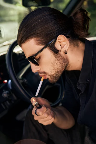 Guapo sexy modelo masculino con pendiente y gafas de sol encendiendo su cigarrillo en el coche, conductor sexy - foto de stock