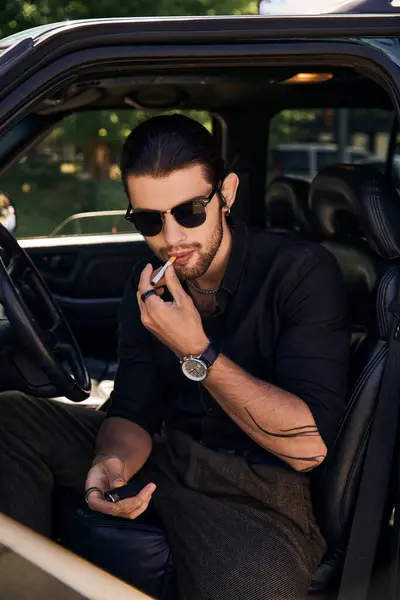 Atractivo modelo masculino joven con reloj de pulsera y tatuaje en elegante traje negro posando con cigarrillo - foto de stock
