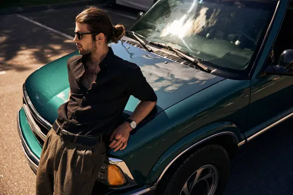 Encantador joven con elegantes accesorios apoyados ligeramente en su coche y mirando hacia otro lado, la moda - foto de stock