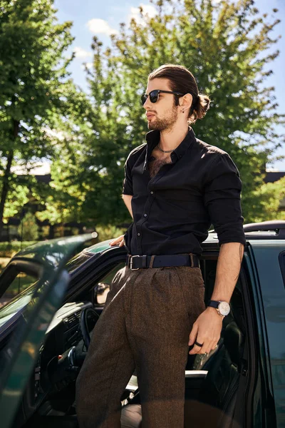 Joven conductor sexy en elegante traje negro con reloj de pulsera y pendiente posando junto a su coche - foto de stock