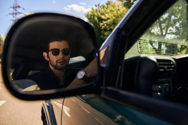 Tentant homme élégant avec des lunettes de soleil en tenue élégante noire regardant dans le miroir latéral, chauffeur sexy — Photo de stock