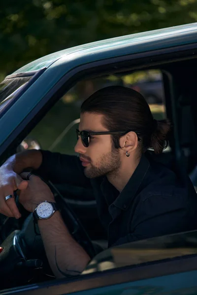 Good looking elegant man with earring and beard in black attire behind steering wheel, sexy driver — Stock Photo