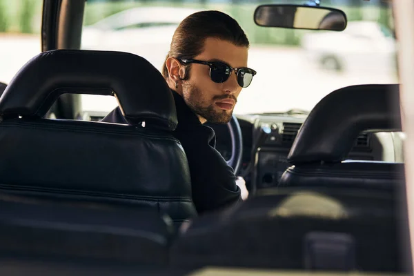 Gut aussehender Fahrer im schwarzen Outfit hinter Lenkrad und Blick in die Kamera, Modekonzept — Stockfoto