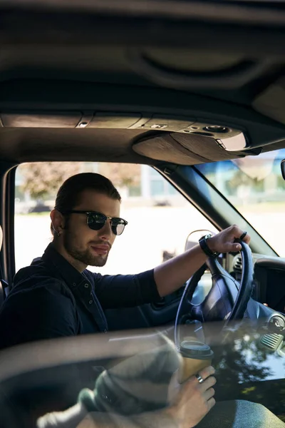 Vertical shot of attractive elegant man with sunglasses and wristwatch at steering wheel with coffee — Stock Photo