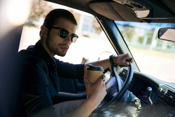 Attraktiver Mann in schwarzer Kleidung hält Lenkrad mit Kaffeetasse in der Hand und blickt in die Kamera — Stockfoto