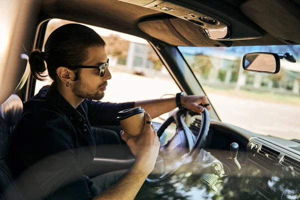 Gut aussehender Mann mit Pferdeschwanz hinter dem Lenkrad seines Autos mit Kaffee in der Hand, sexy Fahrerin — Stockfoto