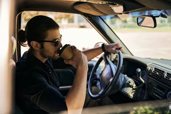 Good looking stylish male model in black outfit drinking coffee behind steering wheel, sexy driver — Stock Photo