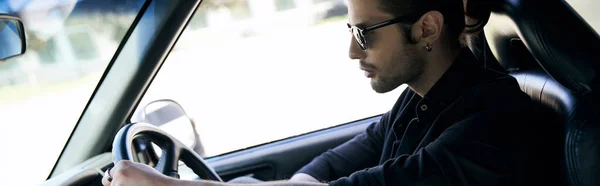Appealing man with accessories in black attire behind steering wheel of his car, sexy driver, banner — стоковое фото
