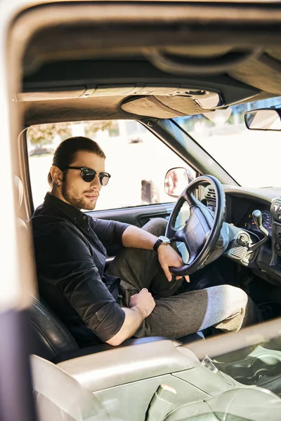 Vertical shot of good looking sexy male model in stylish attire looking at camera at steering wheel — Stock Photo