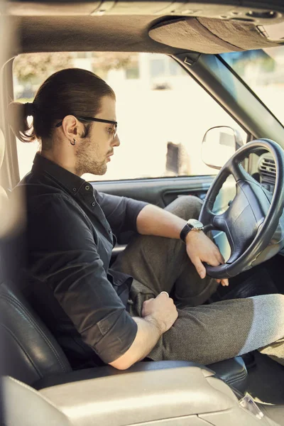 Coûteux modèle masculin élégant avec accessoires en tenue noire derrière le volant, chauffeur sexy — Photo de stock