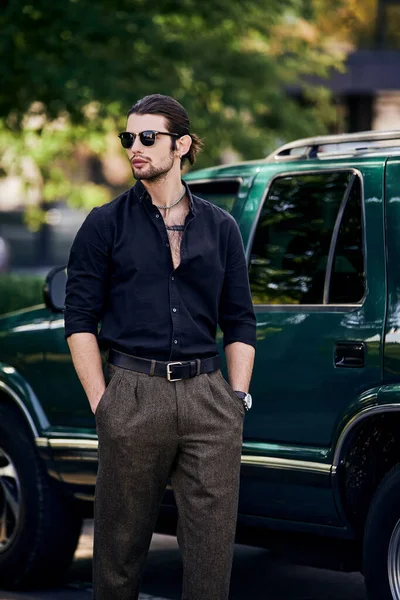 Сексуальна людина з аксесуарами в стильному вбранні, що стоїть поруч зі своєю машиною з руками в кишенях — стокове фото