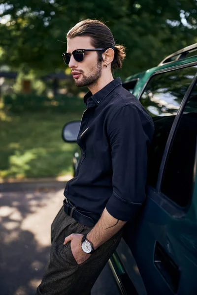 Tiro vertical de un joven guapo en camisa negra con cola de caballo posando cerca del coche, conductor sexy - foto de stock