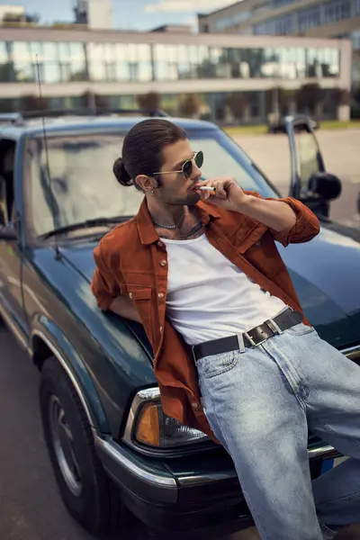 Bel homme sexy en tenue vibrante avec des lunettes de soleil fumant cigarette appuyé sur sa voiture — Photo de stock