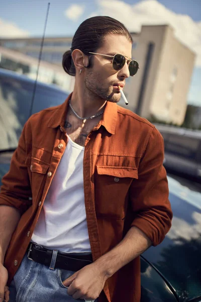 Вертикальный снимок красивого молодого человека в ярком одеянии, позирующего с сигаретой рядом со своей машиной — стоковое фото