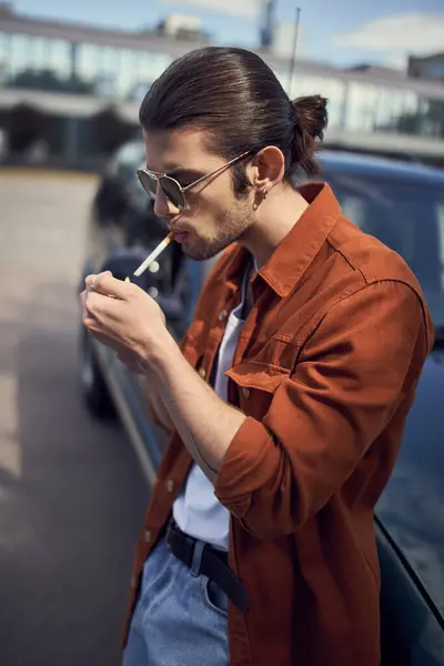Hübsches junges männliches Model mit Pferdeschwanz und Sonnenbrille in braunem Hemd, das seine Zigarette anzündet — Stockfoto