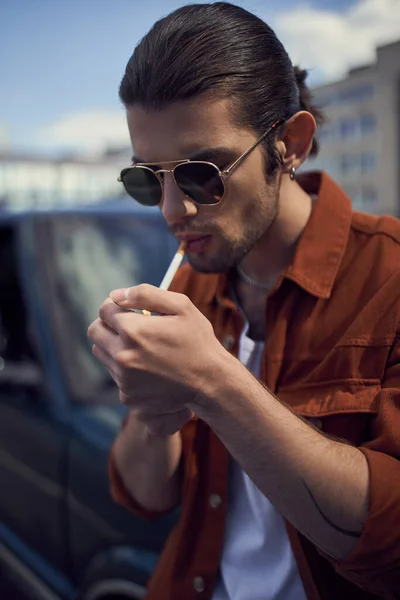 Retrato de un hombre joven y elegante con gafas de sol iluminando su cigarrillo, vertical, concepto de estilo - foto de stock