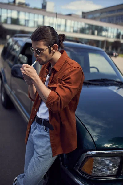 Bello uomo alla moda in giacca marrone e jeans accendendo la sigaretta accanto alla sua auto — Foto stock