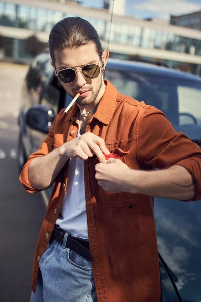 Appealing young man with ponytail and sunglasses posing next to his car with cigarette in his mouth — Stock Photo