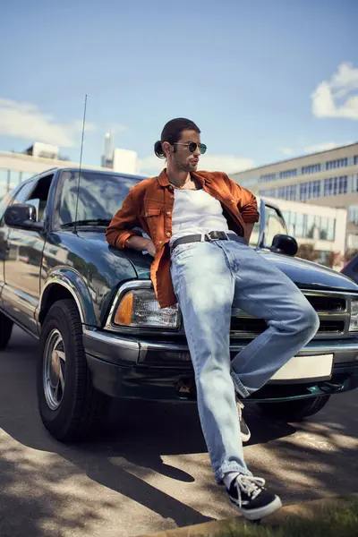 Homem sexy de boa aparência em roupas elegantes vibrantes inclinando-se em seu carro olhando para longe, moda e estilo — Fotografia de Stock