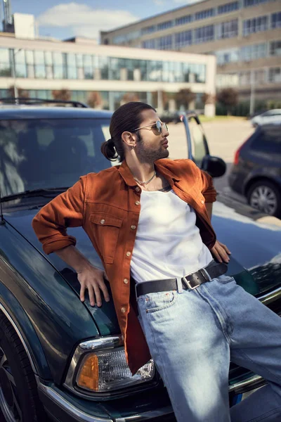 Handsome young man with sunglasses and ponytail in vivid attire leaning on his car, fashion concept — Stock Photo