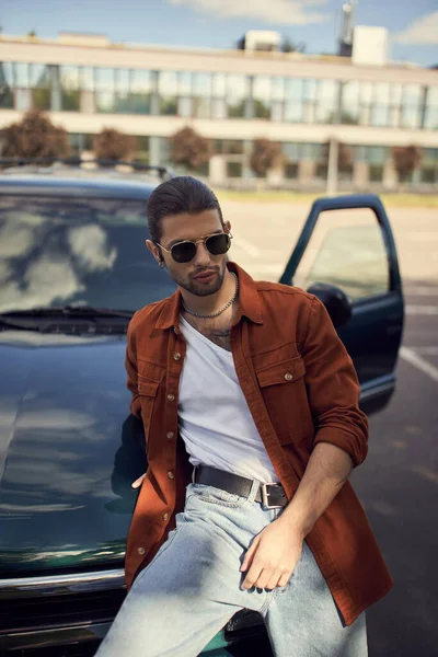 Sexy young man in brown shirt and jeans leaning slightly on his car, fashion and style concept — Stock Photo