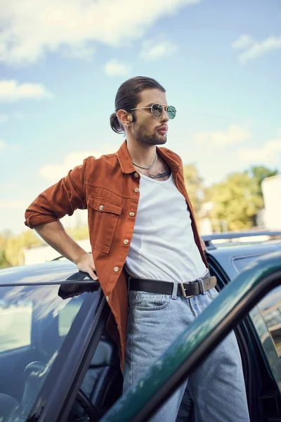 Charmant jeune modèle masculin en chemise marron et jean bleu debout à côté de sa voiture et regardant ailleurs — Photo de stock