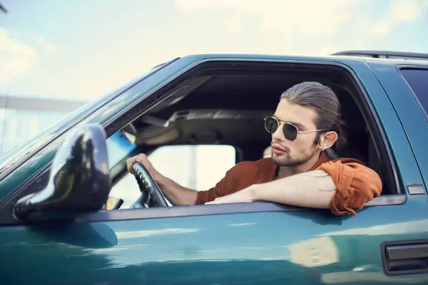 Appealing young man with ponytail and earring in brown shirt looking out of car window, sexy driver — Stock Photo