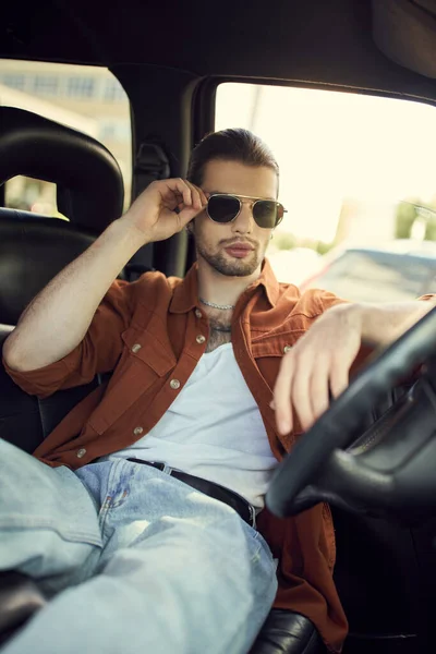 Gut aussehender junger Mann in stylischem Outfit, der seine Sonnenbrille berührt, während er sich im Auto entspannt, sexy Fahrer — Stockfoto