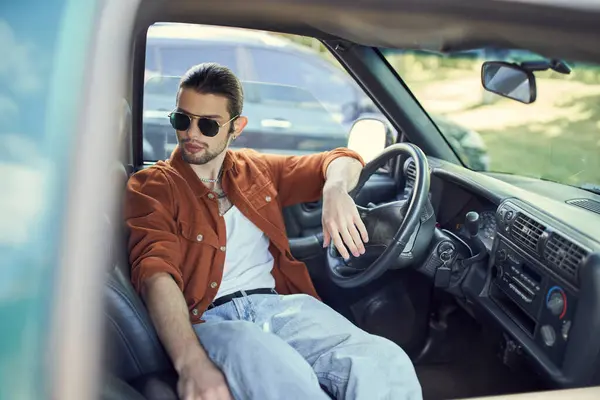 Красивый мужчина в модной коричневой рубашке и джинсах расслабляется в машине за рулем, мода — стоковое фото