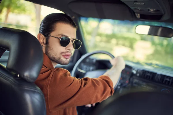 Привабливий стильний чоловік з хвостом і сонцезахисними окулярами позує в його машині і дивиться на камеру — стокове фото