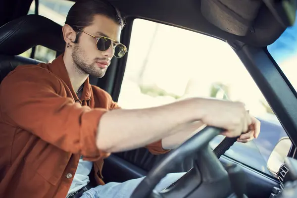 Charming stylish male model in sunglasses with ponytail behind steering wheel of his car, style — Stock Photo