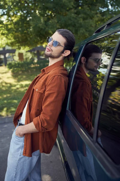 Verführerischer stylischer Mann mit Sonnenbrille, der im Freien posiert, leicht an sein Auto gelehnt, Modekonzept — Stockfoto