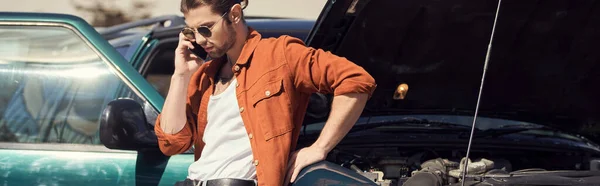 Conductor guapo con gafas de sol en camisa marrón de pie junto a su coche llamando a su aseguradora, pancarta - foto de stock