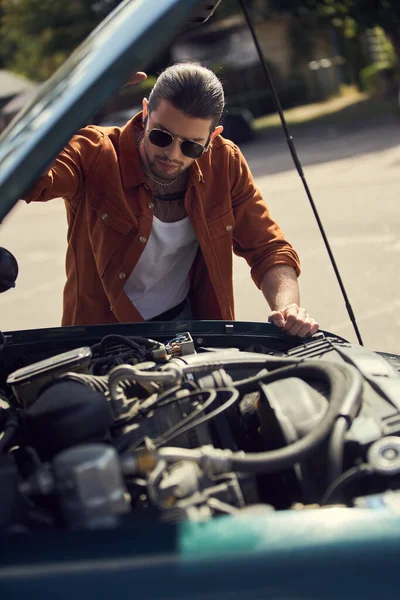 Attrayant jeune homme en chemise marron avec accessoires regardant attentivement le moteur de sa voiture — Photo de stock
