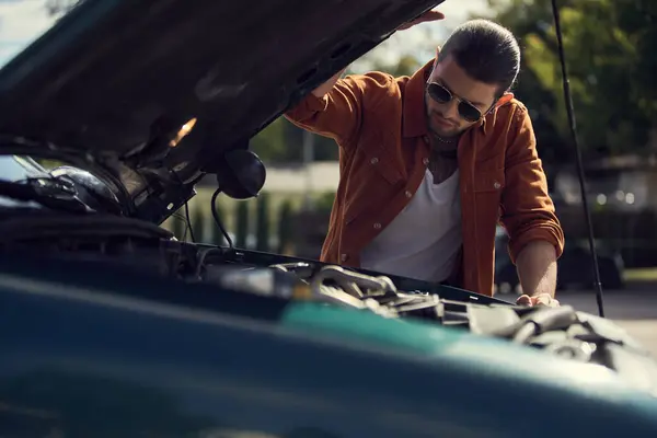 Pensive stylish male model with ponytail and trendy accessories looking at engine of his car — Stock Photo