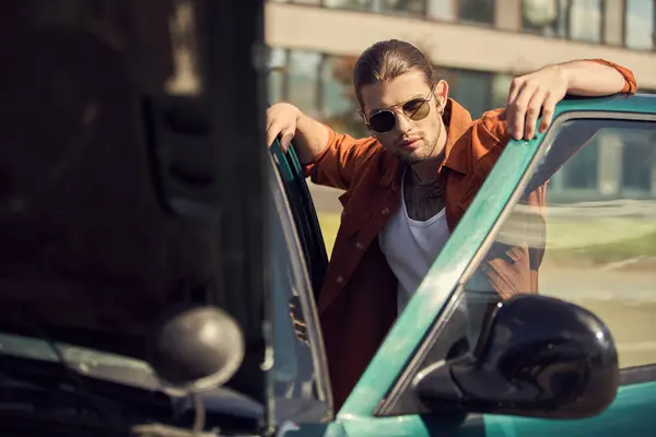 Красивий сексуальний водій у коричневій сорочці з сонцезахисними окулярами дивиться на камеру, позуючи поруч зі своєю машиною — стокове фото